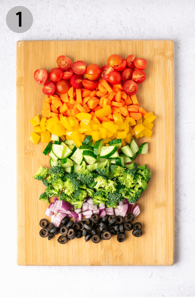 chopped veggies on a wood board: tomatoes, carrots, peppers, cucumber, broccoli, red onion, black olives