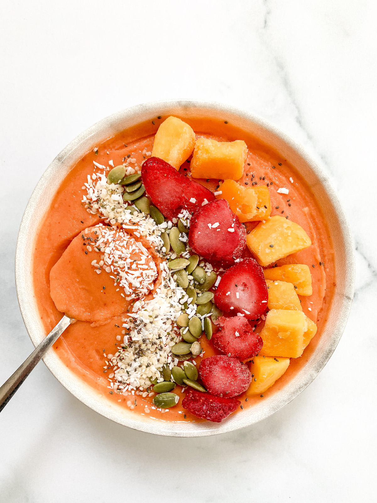 a mango smoothie bowl topped with coconut flakes, pumpkin seeds, strawberries, and mango pieces with a spoon in it.