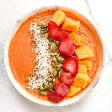 a mango smoothie bowl topped with coconut flakes, pumpkin seeds, strawberries, and mango pieces.