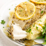 close up of fork flaking a halibut filet with lemon slices on top.