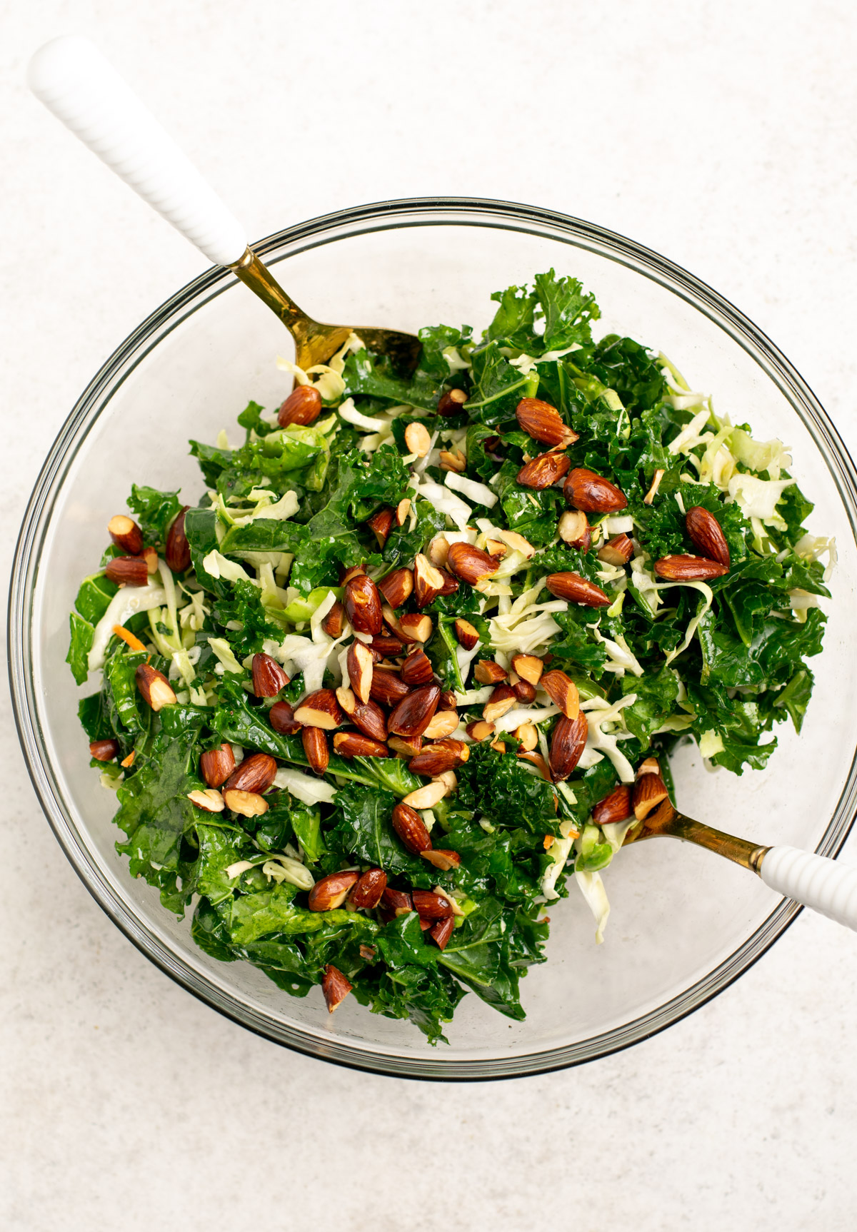 top view of a kale salad in a glass bowl with almonds on top and gold salad spoons.