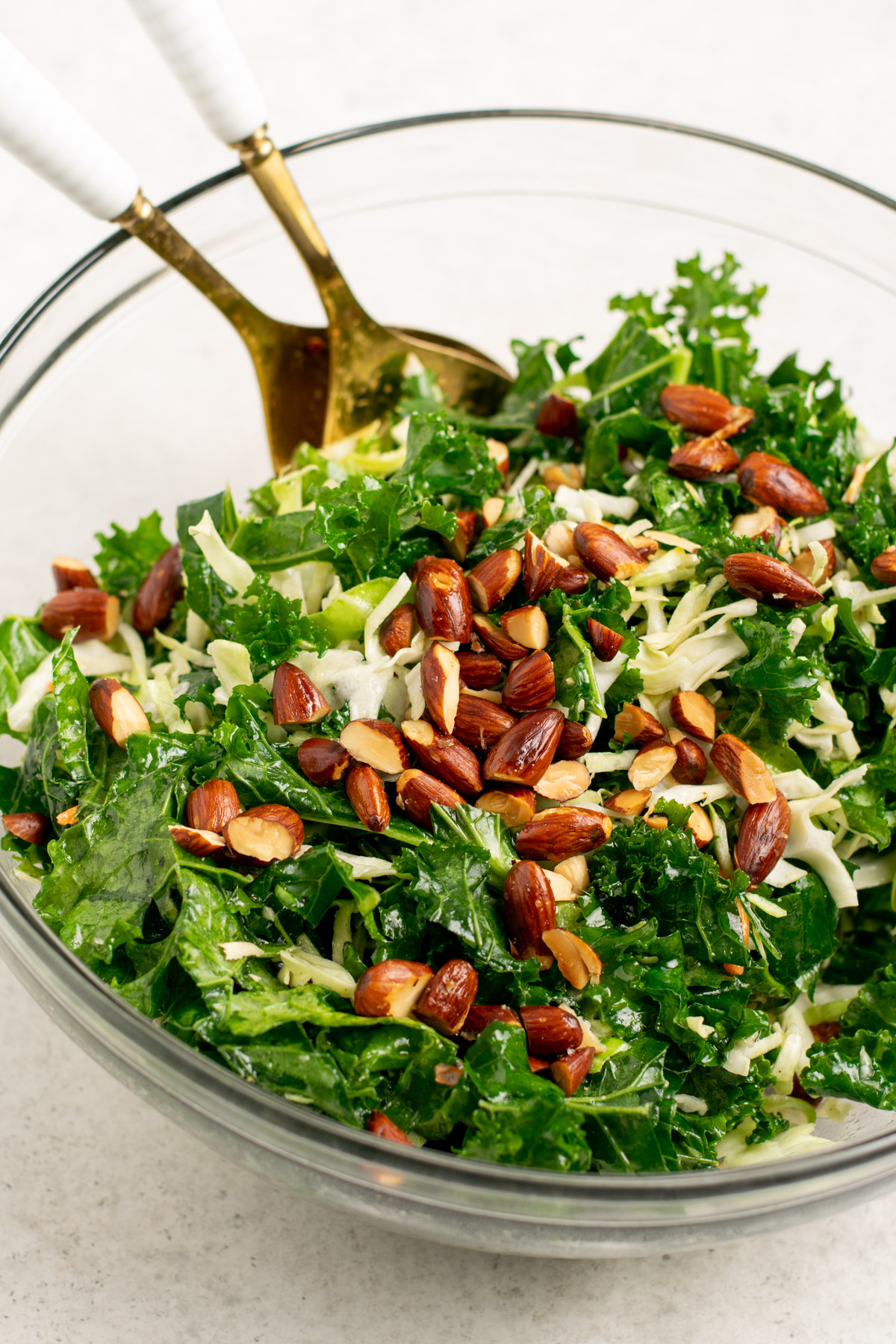 kale salad with almonds on top and gold salad spoons in a glass bowl.