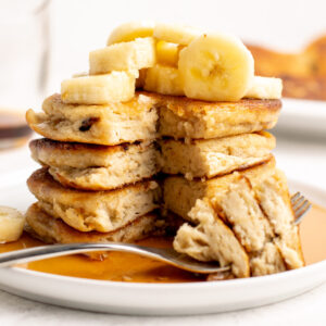 a stack of 4 vegan pancakes covered in syrup with bananas on top. A fork is laying down with a bite on it.