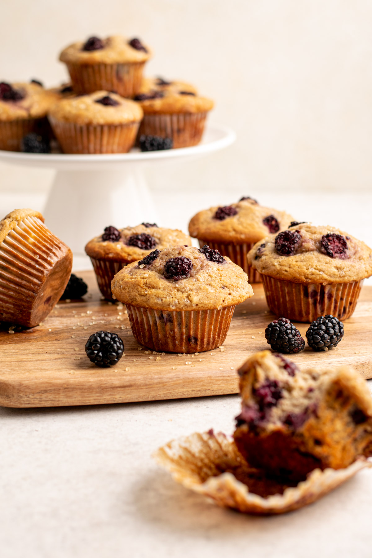 gluten free blackberry muffins on a wood board, with more on a white pedestal behind.