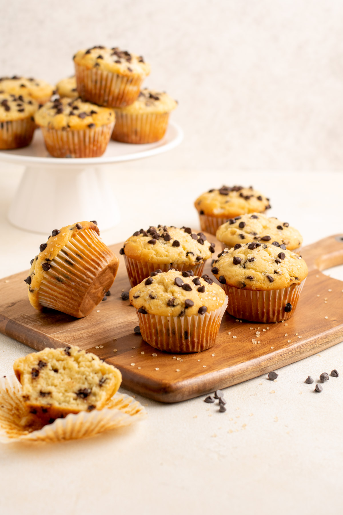 chocolate chip muffins on a wood board, some behind on a white pedestal.