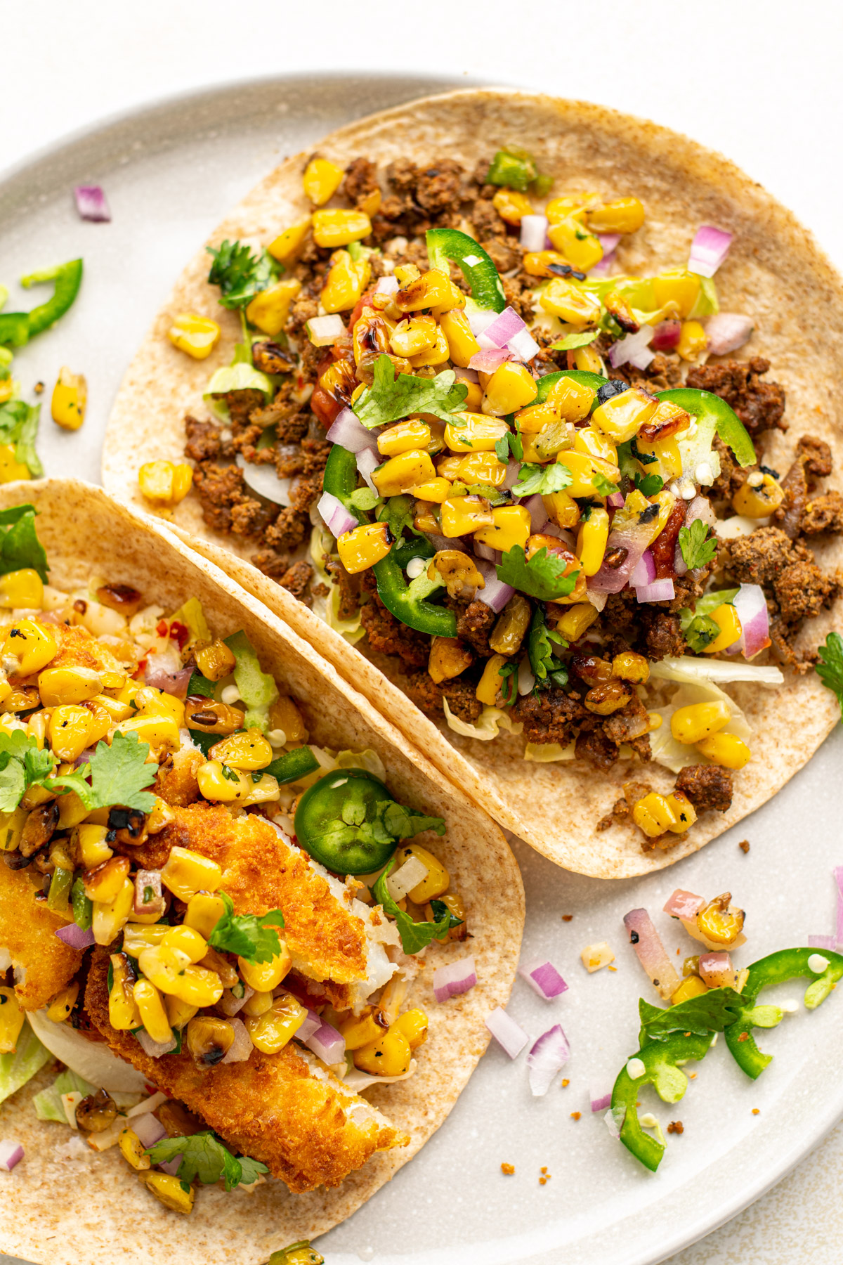 2 tacos on a plate loaded with jalapenos, corn, beef, and cilantro.