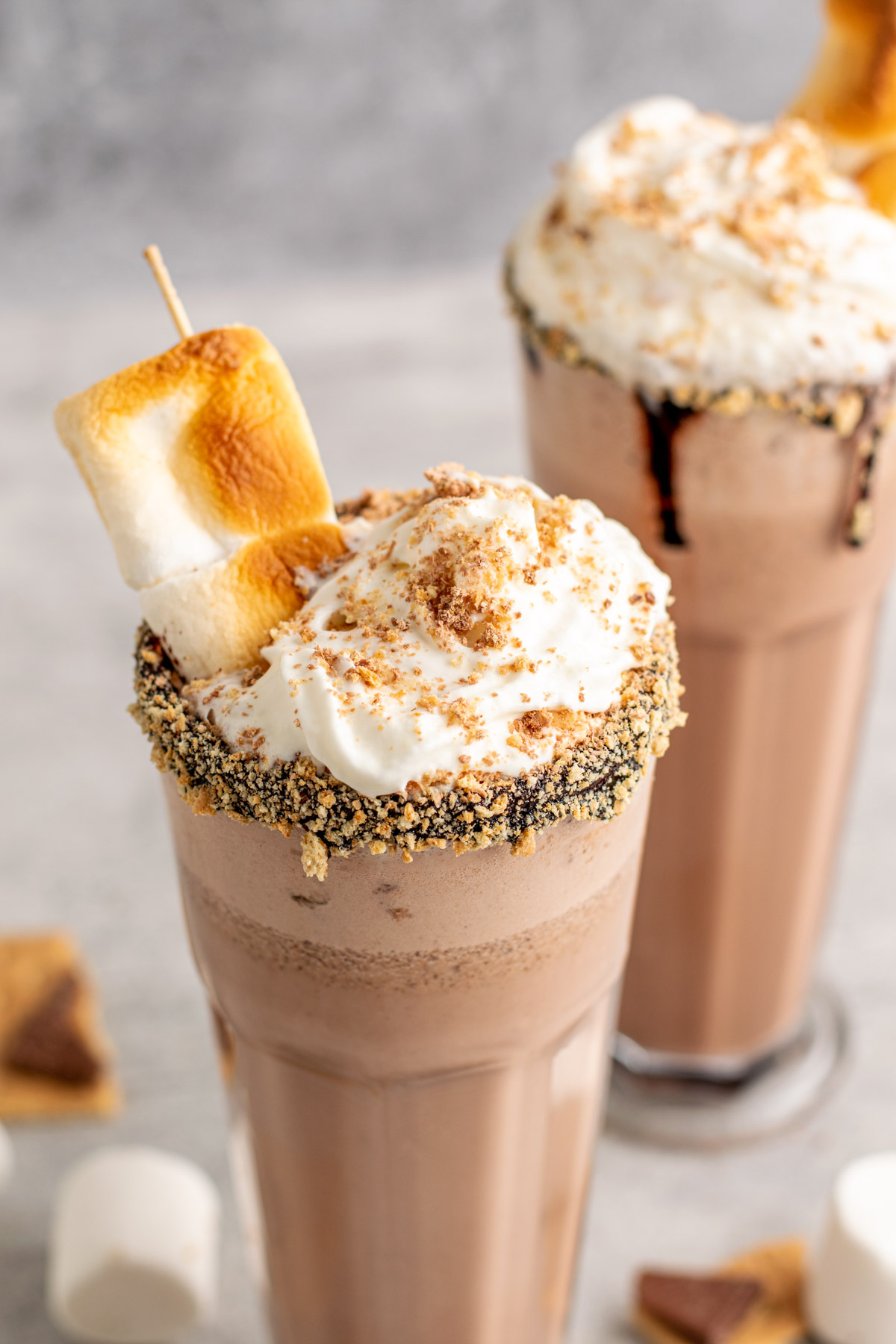 two s'mores milkshakes topped with whipped cream, graham crumbs, and toasted marshmallows.