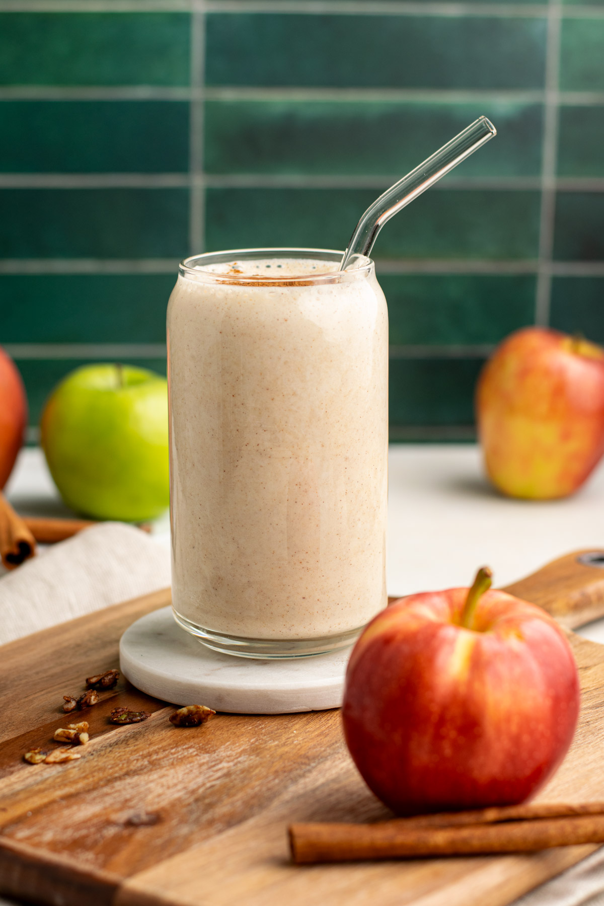 apple pie smoothie in a glass with a glass straw and apples around with a green tile background.