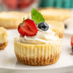 close up of a mini cheesecake topped with whipped cream, half a strawberry, a blueberry, and mint.