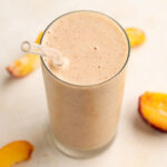 peach smoothie on a tall glass with a glass straw and peach slices around.