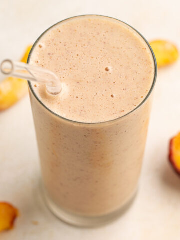 peach smoothie on a tall glass with a glass straw and peach slices around.