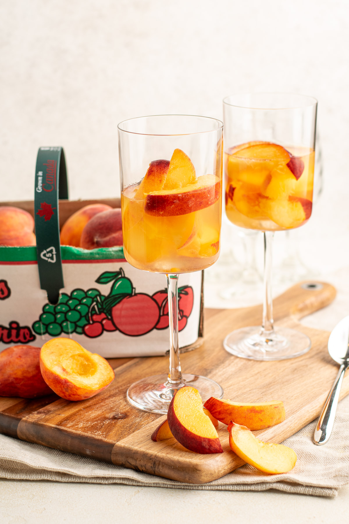 two wine glasses with peach slices and wine, and a basked with peaches in the back.