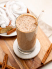 cinnamon roll smoothie in a tall glass with a glass straw and cinnamon buns in the background.
