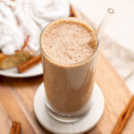 cinnamon roll smoothie in a tall glass with a glass straw and cinnamon buns in the background.