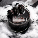 black margarita in a glass with 3 blackberries on top surrounded by spiderwebs.