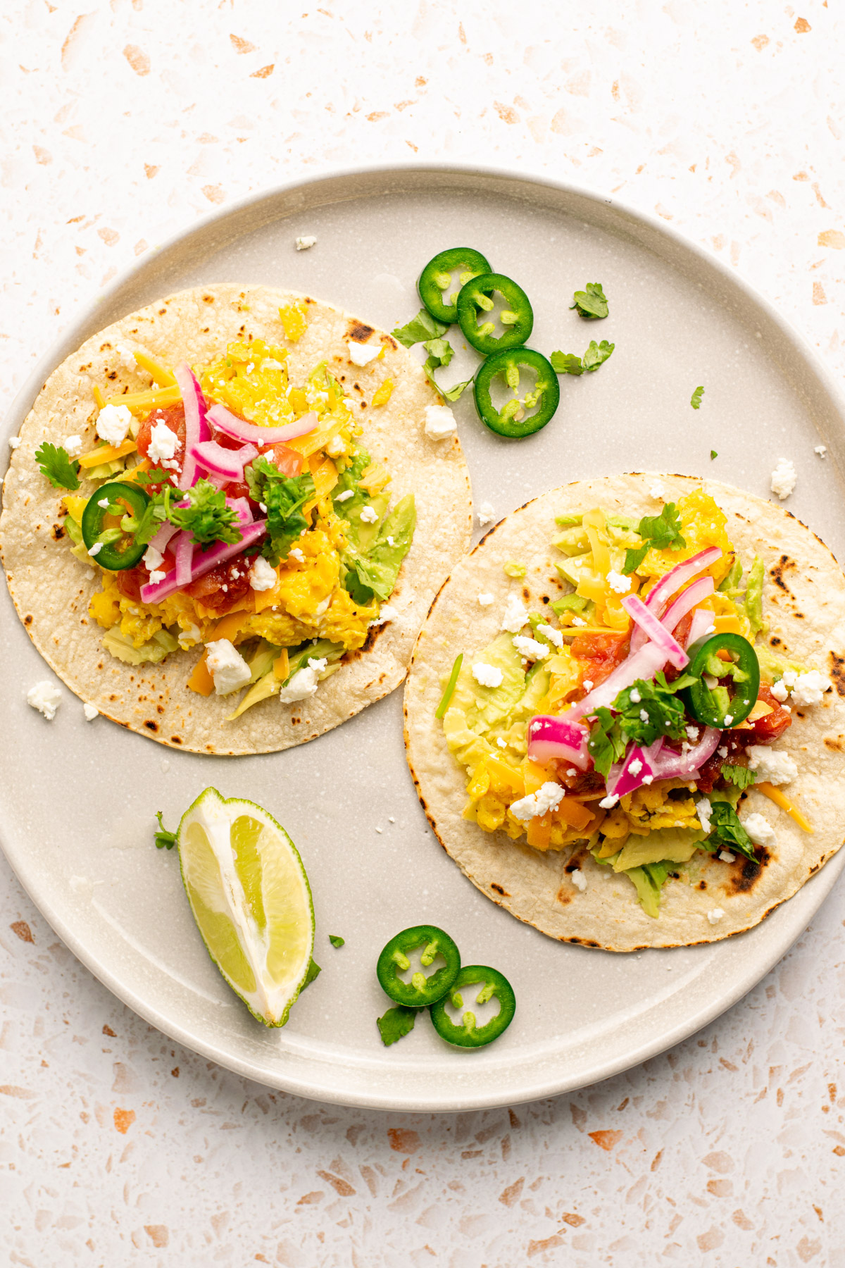 2 breakfast tacos on a plate topped with pickled red onions, cilantro, and jalapeno slices.