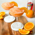 two blended aperol spritz cocktails in coupe glasses with half orange slices.