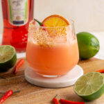 aperol coconut margarita with limes and red chilis around and aperol bottle in the background.